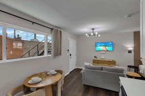 Uma sala de estar moderna com área de jantar, um sofá cinza, TV fixada na parede e uma escadaria visível pela janela.