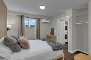 Ein Schlafzimmer mit Bett, Nachttisch, Klimaanlage, einem Fenster mit Vorhängen, einer Waschmaschine und einem Kleiderschrank.