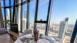 A dining area with a table set for two, a view of a cityscape through large windows, and a plant on the left. - Best for get away & Peaceful Haven by Flouri F282O (Seef)
