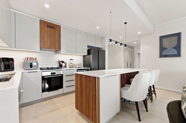 A modern kitchen with white cabinetry, a built-in oven, a stove, and a dining area with a white island and two chairs. - Luxury 3 Bed Apartment |Balcony, Pool and Gym (Umhlanga)