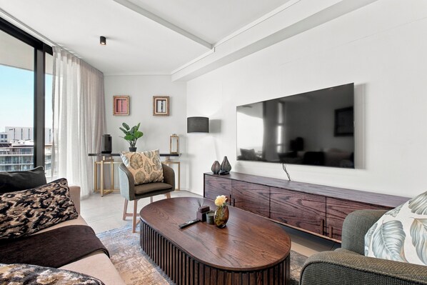 A modern living room with a flat-screen TV, a sofa, a coffee table, and a view of the city. - Luxury 3 Bed Apartment |Balcony, Pool and Gym (Umhlanga)