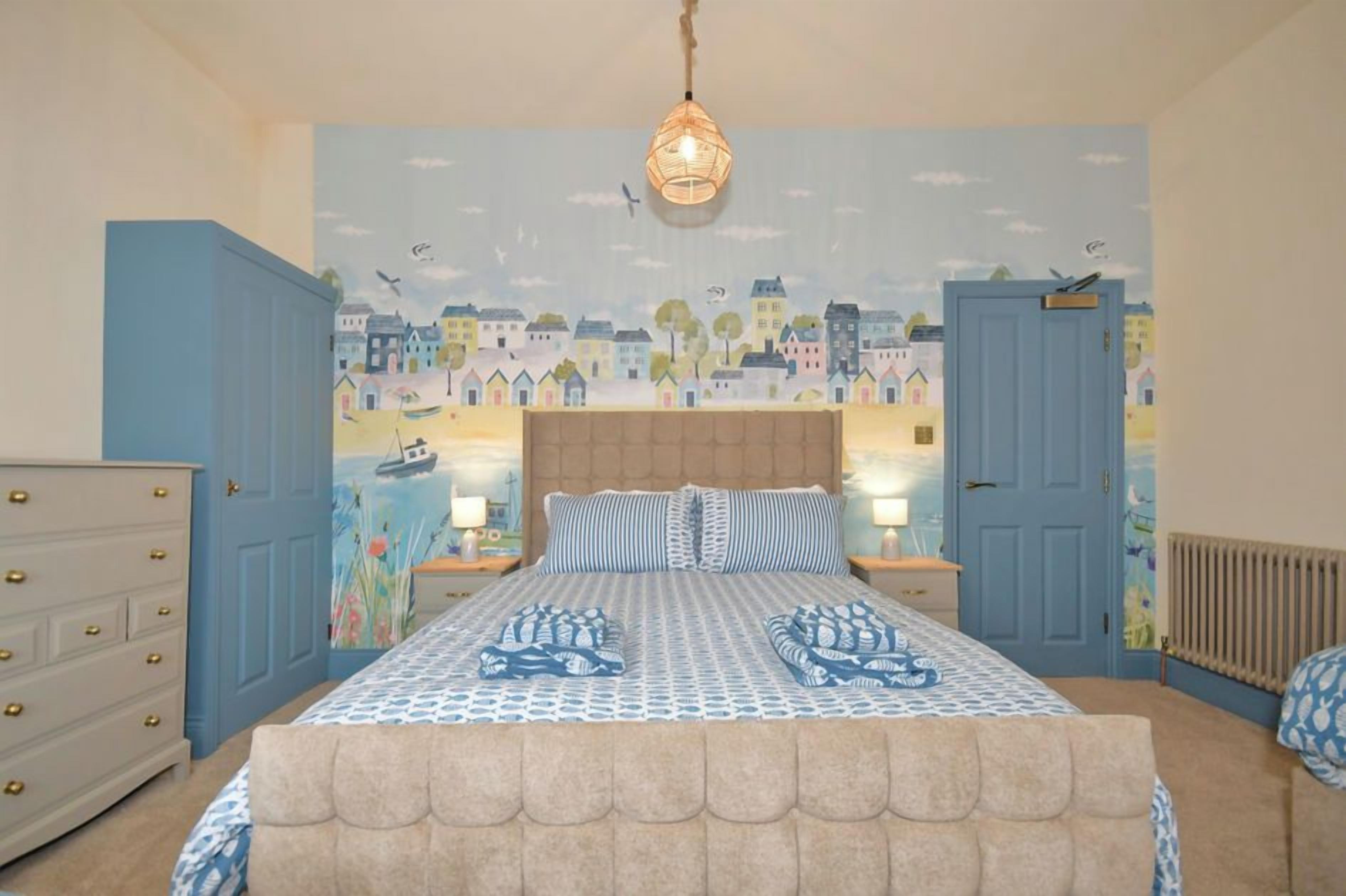 A bedroom with a bed, two bedside tables, a dresser, a wall mural, and a hanging light fixture.