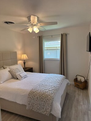 Ein Schlafzimmer mit Bett, Nachttisch, Lampe, Deckenventilator, Fenster mit Vorhängen und einem an der Wand montierten Fernseher.