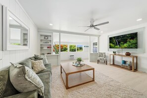 A modern living room with a sofa, coffee table, and TV. There are shelves with books and decorative items, and a large window with a view. - Kahaluu Beach, 2-bed 2-bath, AC, steps to beach (Kailua-Kona)