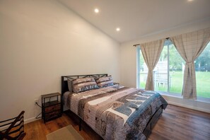 A bedroom with a bed, a bedside table, a nightstand, a window with curtains, and a view of the outdoors. - Lakefront Champlain Duplex , King Bed & Bunk Bed w/ Kayaks, Pool & Fire Pit (Champlain)