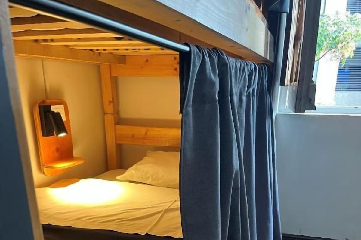 Una cama litera con una lámpara colgante, una ventana con cortinas y piso de madera.