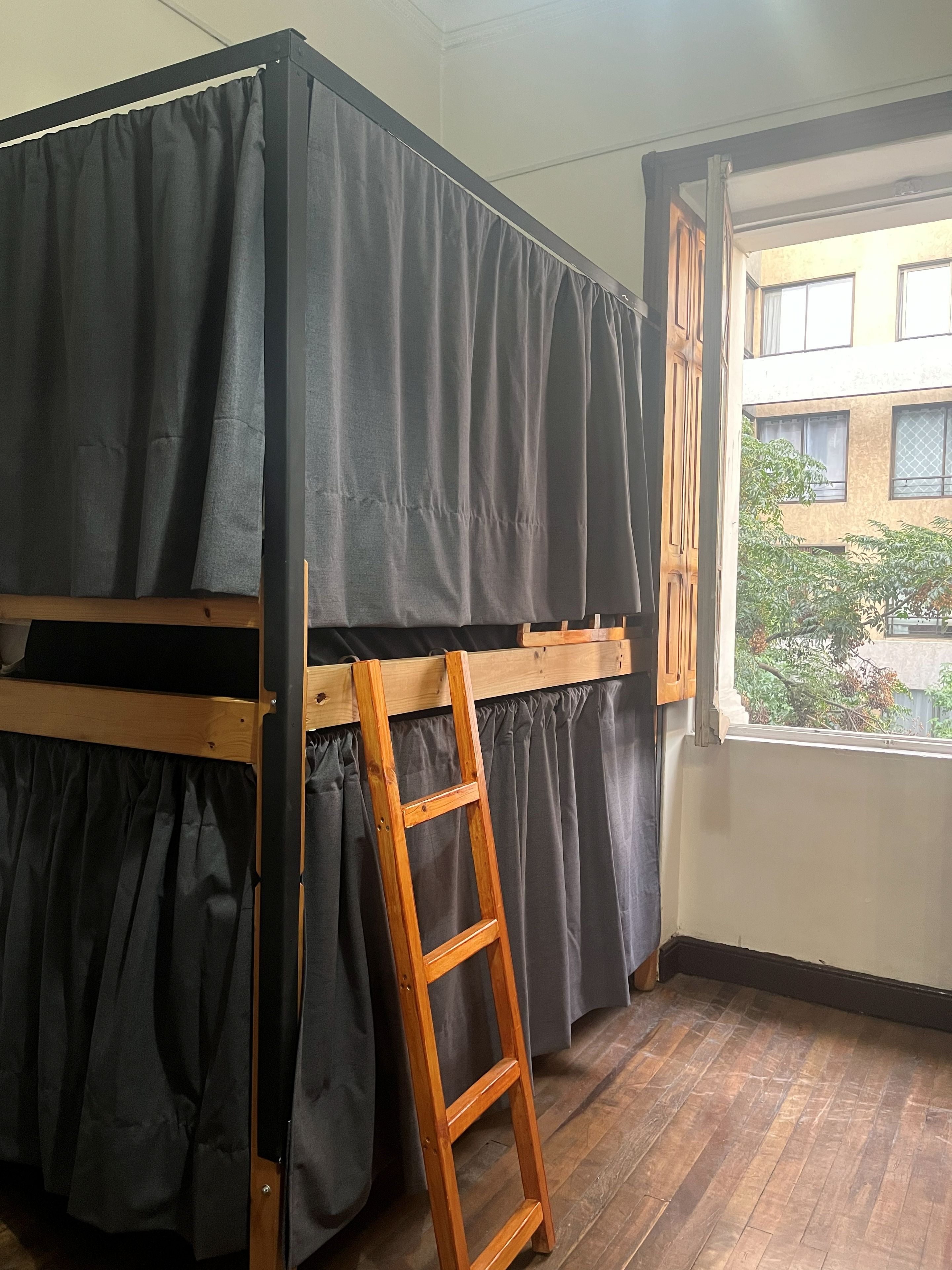 Una cama nido con escalera de madera, cortinas negras y una ventana con vistas a edificios y árboles.