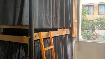 Una cama nido con escalera de madera, cortinas negras y una ventana con vistas a edificios y árboles.