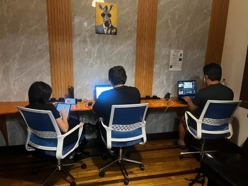 Tres personas sentadas en un escritorio de madera con computadoras, en una habitación con pared de mármol y una obra de arte con motivos de cebra.