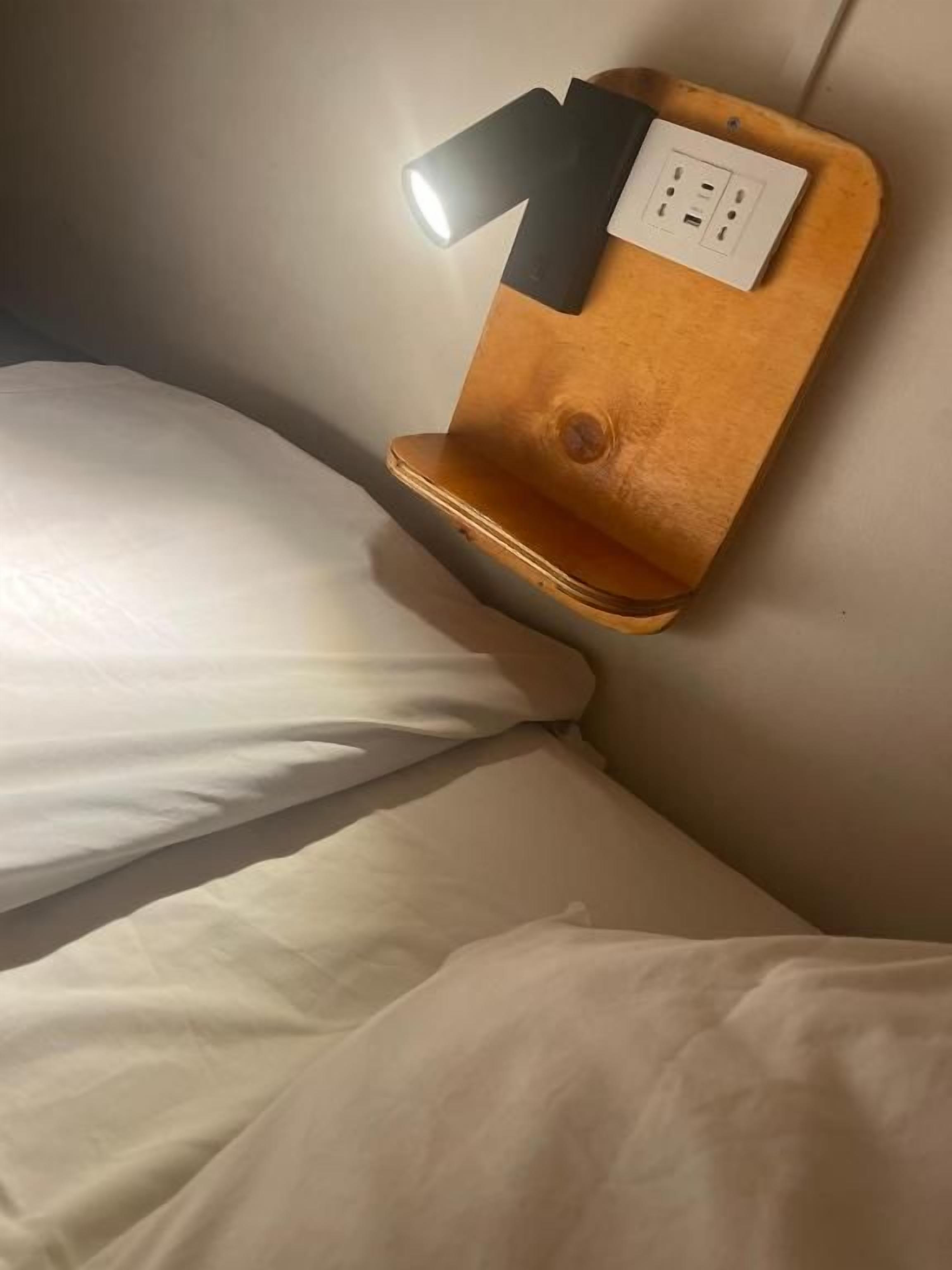 Una cama con ropa de cama blanca, una mesita de noche fijada a la pared con una lámpara y un enchufe.