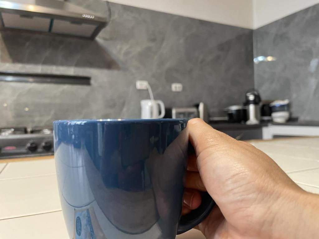 Una mano sosteniendo una taza azul en una cocina de diseño moderno, con una estufa y una cafetera.