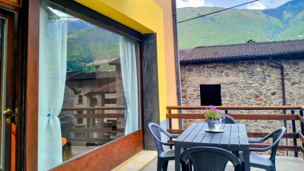 A balcony with a table and chairs, a window with curtains, and a view of a mountain. - Vrbo Property (Forcola)