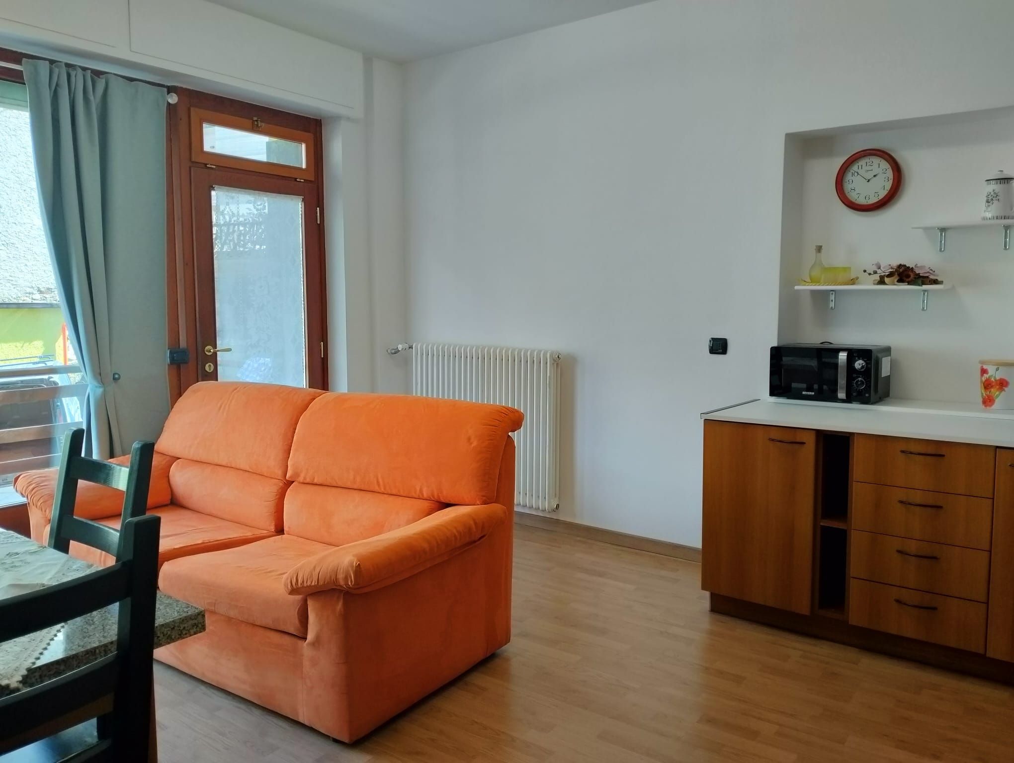 En stue med sofa og spisebord, et køkken med mikroovn, og et vindue med gardiner.