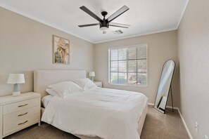 Ein Schlafzimmer mit Bett, Nachttischen, Deckenventilator, Fenster mit Jalousien und Spiegel.