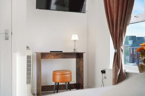 A hotel room with a wooden console table, a single stool, a wall-mounted TV, a bedside lamp, and a window with curtains. - Serene 2BR House | Garden & Parking | Stay With Us (Stockton-on-Tees)