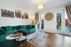 Un salon avec un canapé vert, une petite table ronde sur laquelle se trouvent un livre et une télécommande, une grande horloge murale et une fenêtre avec des rideaux.