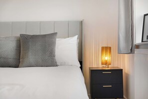 A neatly made bed with two pillows, a bedside table with a lamp, and a radiator. - Serene 2BR House | Garden & Parking | Stay With Us (Stockton-on-Tees)