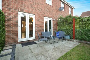 A brick house with a patio area featuring a metal table and chairs, a satellite dish, and a green hedge. - Serene 2BR House | Garden & Parking | Stay With Us (Stockton-on-Tees)