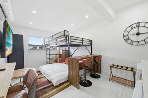Ein kleines Zimmer mit Stockbetten, einem Schreibtisch, einem Stuhl, einer Uhr und einem Fenster.