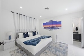 Un dormitorio moderno con una cama grande, una mesita de noche, un radiador fijado a la pared y un cuadro con un atardecer sobre un puente.