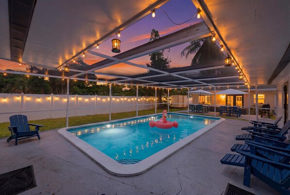 Un área de piscina al aire libre con un patio cubierto, luces colgantes y sillas de descanso.