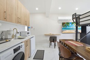 A compact kitchen with modern appliances, a dining area with a wooden table, and a beach-themed wall art. - 10min Hard Rock - Studio, Patio, Workspace (Miami Gardens)