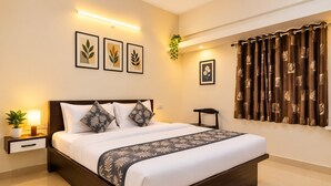 Quarto de hotel com uma cama grande, mesa de cabeceira com abajur, uma cadeira pequena, planta em vaso e uma janela com cortinas.