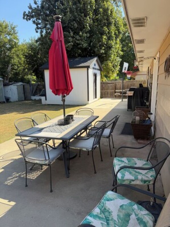 A patio with a table and chairs, a red umbrella, and a small shed in the background.