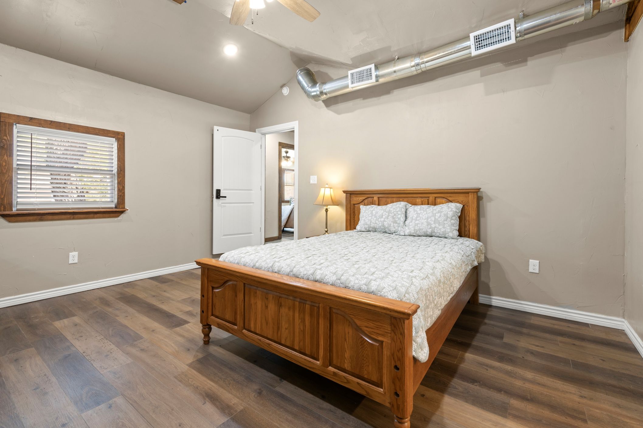 Un dormitorio con cama de madera, ventana con persianas, puerta y rejilla en el techo.
