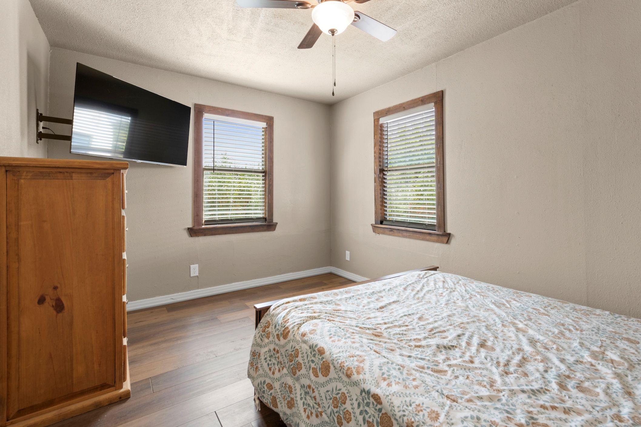 A bedroom with a bed, a wooden wardrobe, a ceiling fan, and two windows with blinds.