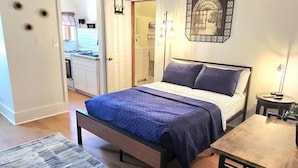 A bedroom with a bed, a wooden table, and a bench. There is a door leading to a kitchen. - The Tidy Third Floor Studio (Buffalo)