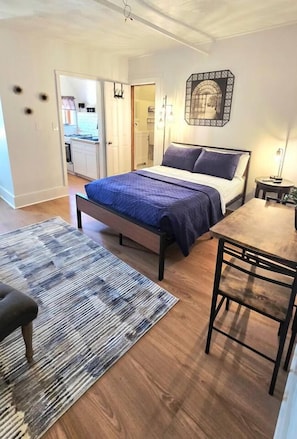 Un dormitorio con cama, una mesa de madera y un banco. Hay una puerta que da a una cocina.