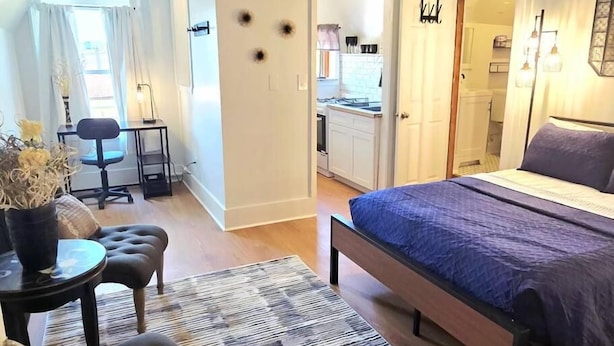 A bedroom with a bed, a desk, a chair, and a small table with a vase. - The Tidy Third Floor Studio (Buffalo)