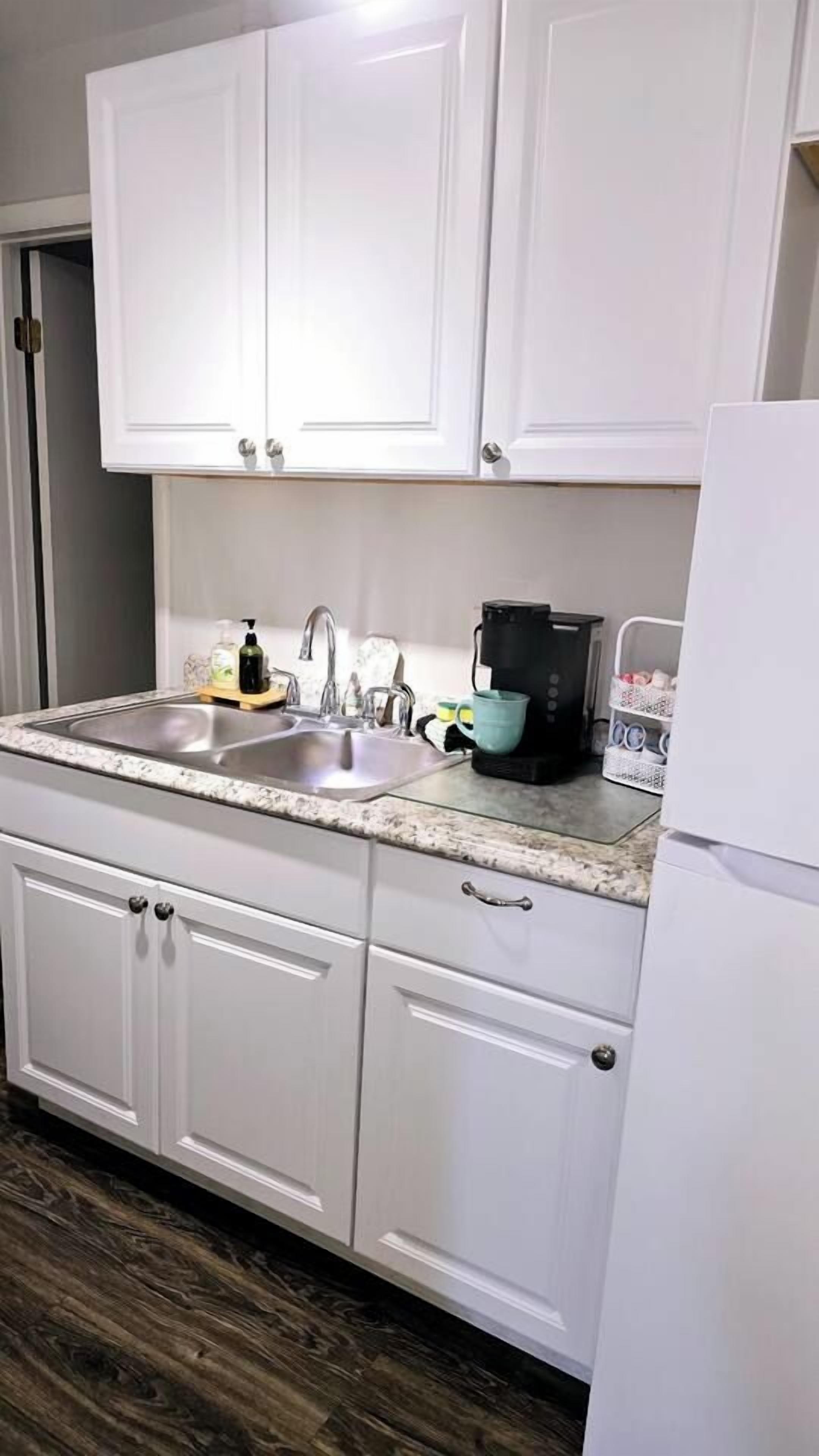Una cocina con armarios blancos, un lavamanos doble, una cafetera y un refrigerador.