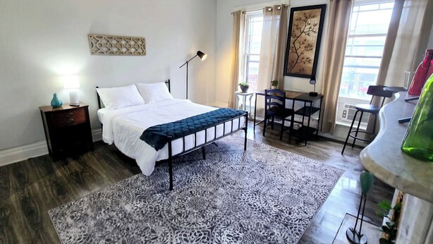 A bedroom with a bed, bedside table, lamp, desk, chair, and a view of the outside. - Cozy Studio in Allentown (Buffalo)