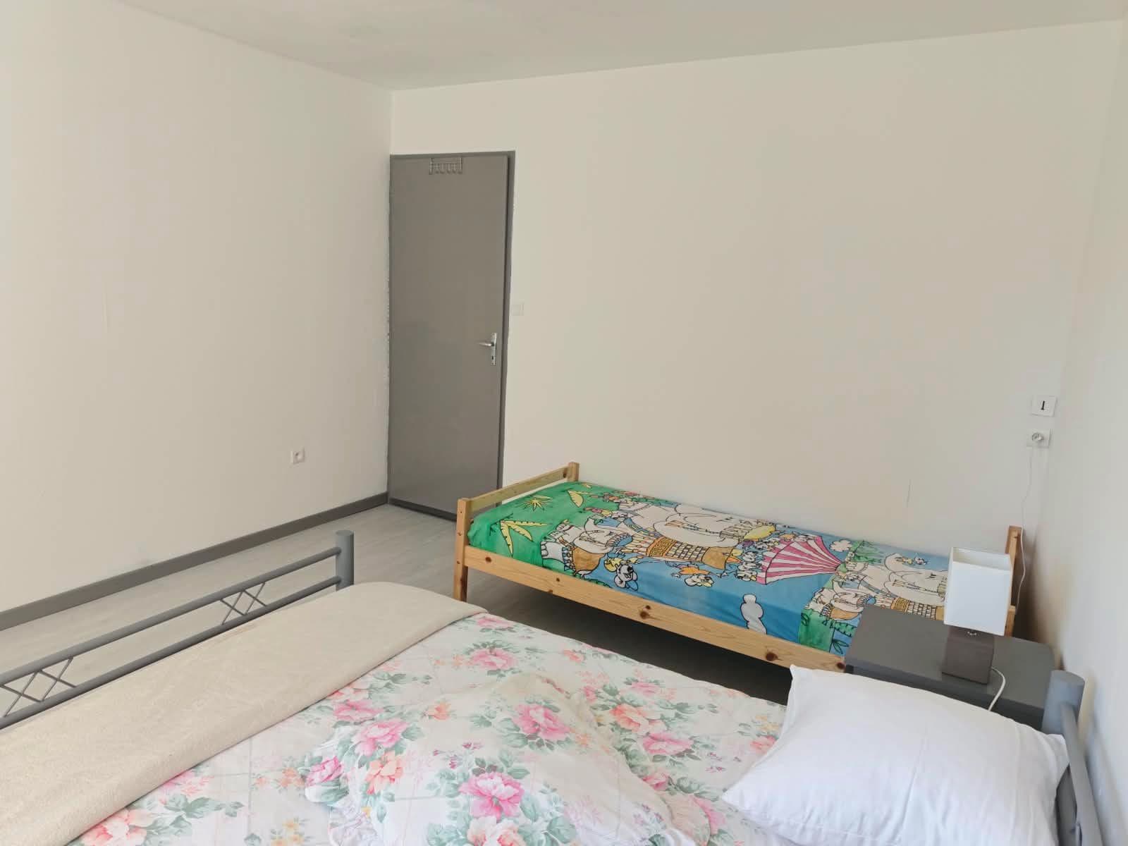 Un lit simple avec une couvre-lit à motifs floraux, une table de chevet avec une lampe, et une porte visible au fond.