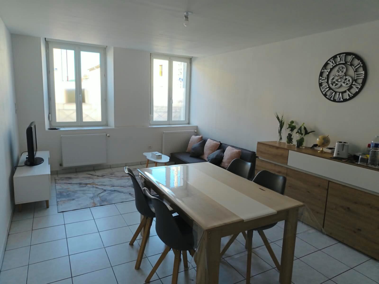 A modern kitchen with a dining table, chairs, and a sofa. There are windows, a clock on the wall, and a radiator.