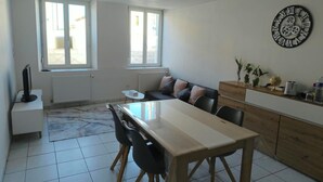 A modern kitchen with a dining table, chairs, and a sofa. There are windows, a clock on the wall, and a radiator.