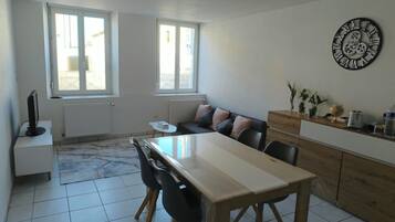 A modern kitchen with a dining table, chairs, and a sofa. There are windows, a clock on the wall, and a radiator.