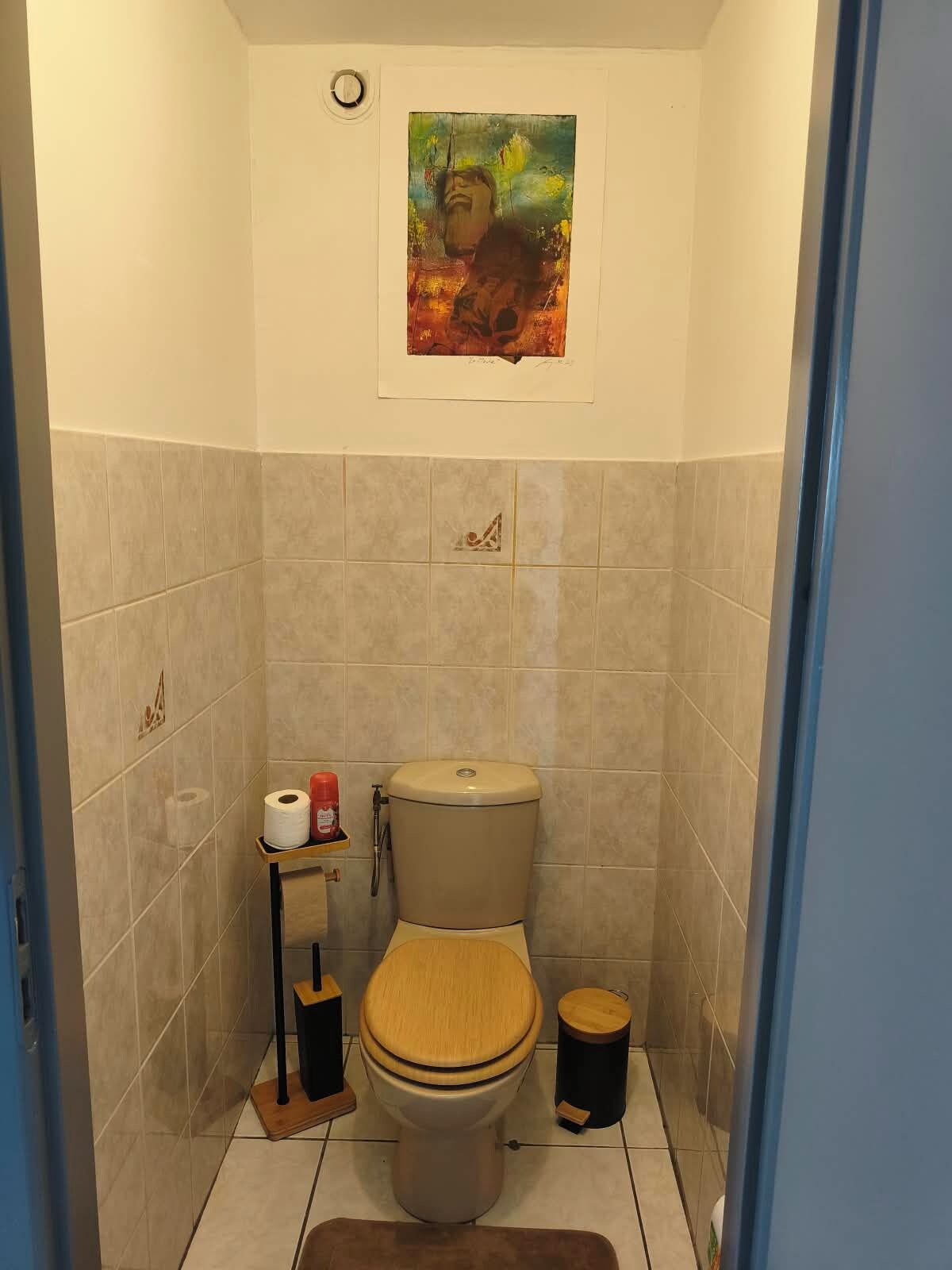 A bathroom with a toilet, a small shelf with toilet paper and a red bottle, a wooden stool, and a painting on the wall.