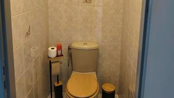 A bathroom with a toilet, a small shelf with toilet paper and a red bottle, a wooden stool, and a painting on the wall.