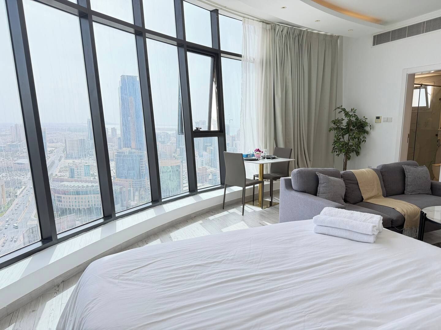 A hotel room with a large bed, a sofa, a dining table with chairs, and a view of the cityscape through floor-to-ceiling windows.