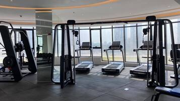 A modern gym with cardio machines, weight training equipment, and large windows offering a view of the city.