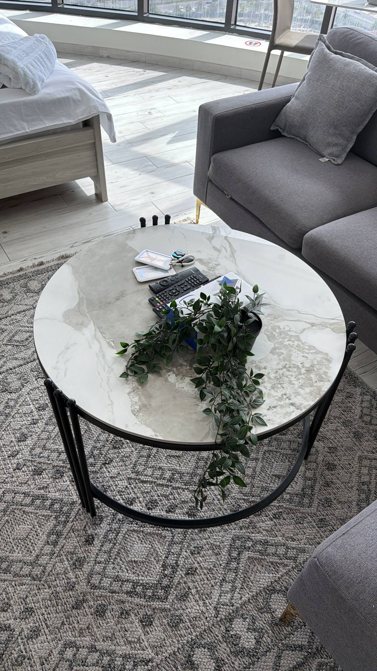 A round coffee table with a marble top, surrounded by a patterned carpet, with a grey sofa and a white bed in the background.