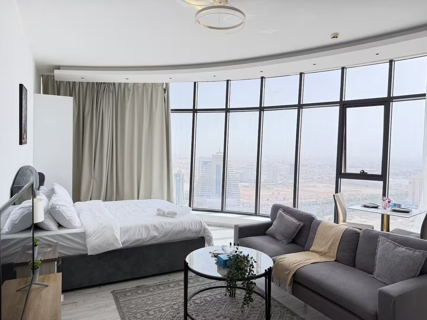 A modern hotel room with a bed, a sofa, a small table, and large windows offering a city view.