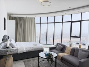 Et moderne hotelværelse med seng, sofa, lille bord og store vinduer med udsigt over byen.