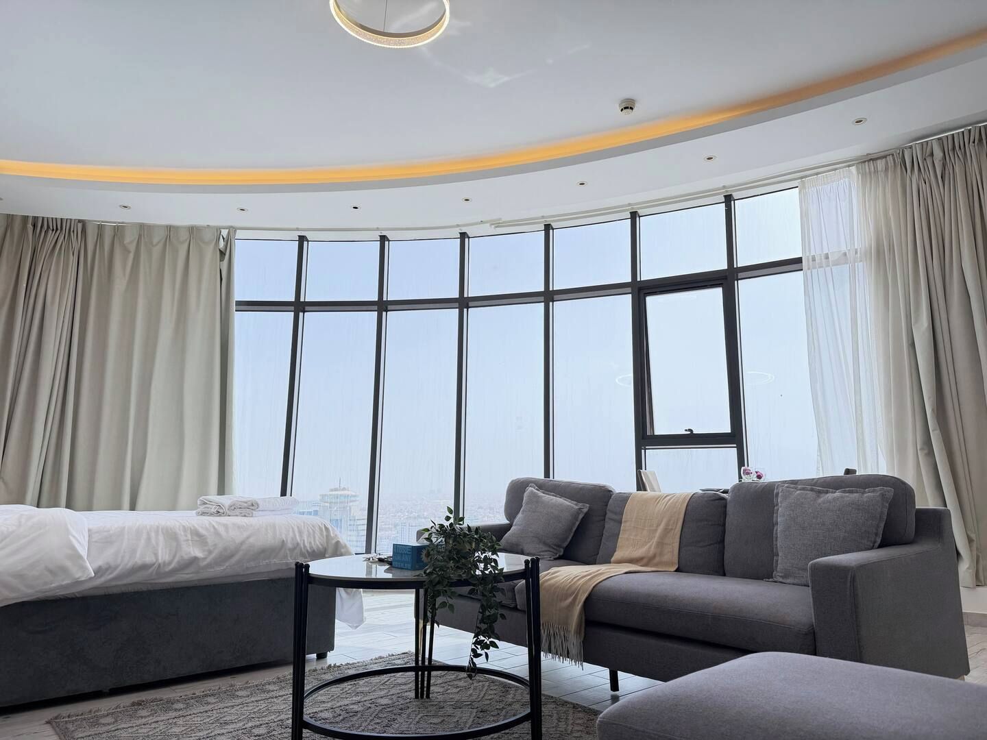 A modern hotel room with a curved ceiling, a large window offering a city view, a bed, a sofa, and a small table with a plant.