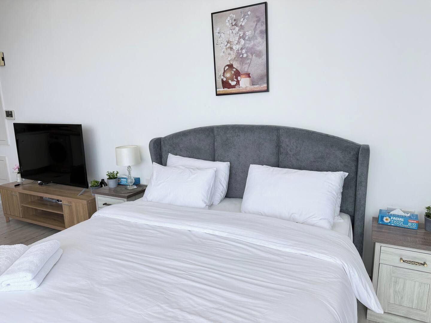 A neatly made bed with white linens, a grey upholstered headboard, a bedside table with a lamp, and a television on a wooden stand.