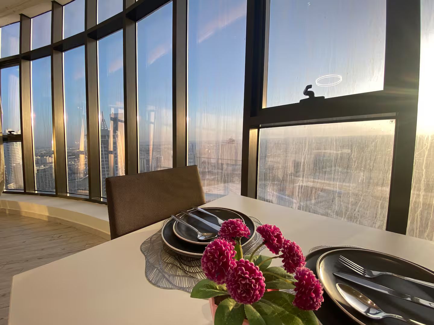A dining area with a table set for two, a chair, and a view of the cityscape through large windows.
