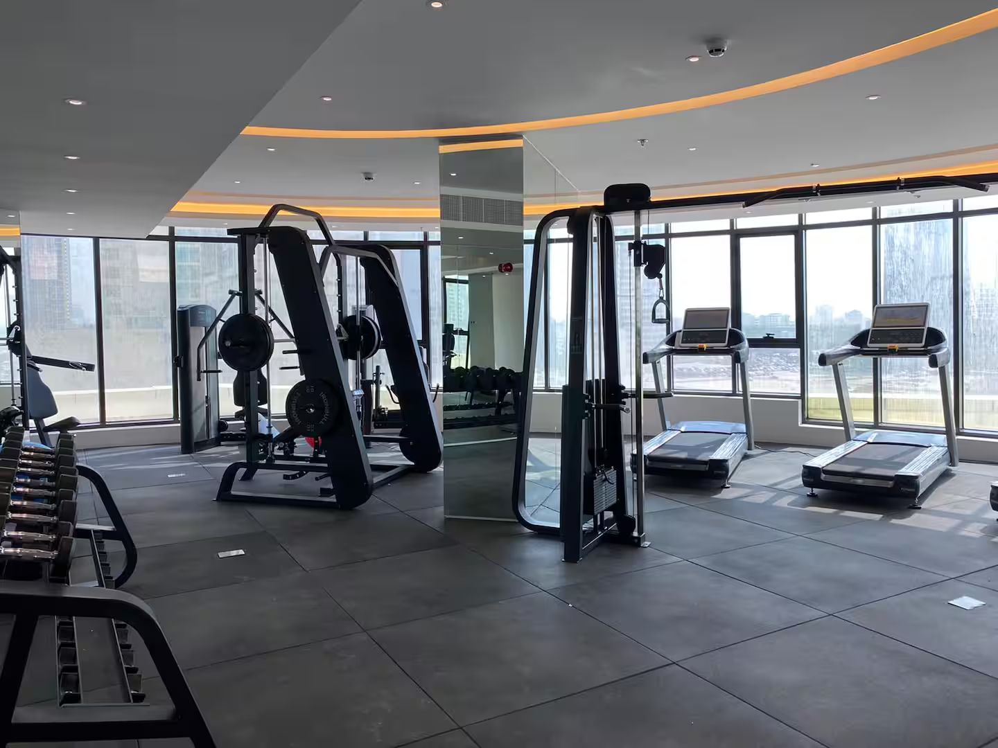 A modern gym with various exercise equipment, large windows, and a view of the cityscape.